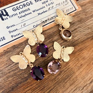 Pu&ograve; includere: Orecchini a farfalla dorati con pendenti di gemme. Gli orecchini presentano farfalle e gemme ovali viola e rosa. &Egrave; visibile anche un'etichetta vintage con il testo "Sapphiresize 8 mm".
