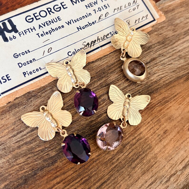 Pu&ograve; includere: Orecchini a farfalla dorati con pendenti di gemme. Gli orecchini presentano farfalle e gemme ovali viola e rosa. &Egrave; visibile anche un'etichetta vintage con il testo "Sapphiresize 8 mm".