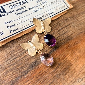 Pu&ograve; includere: Orecchini a farfalla color oro con pietre preziose sfaccettate viola e rosa. Gli orecchini sono esposti su una superficie di legno con un biglietto da visita vintage sullo sfondo. La carta recita "Sapphire Size 18 MM".