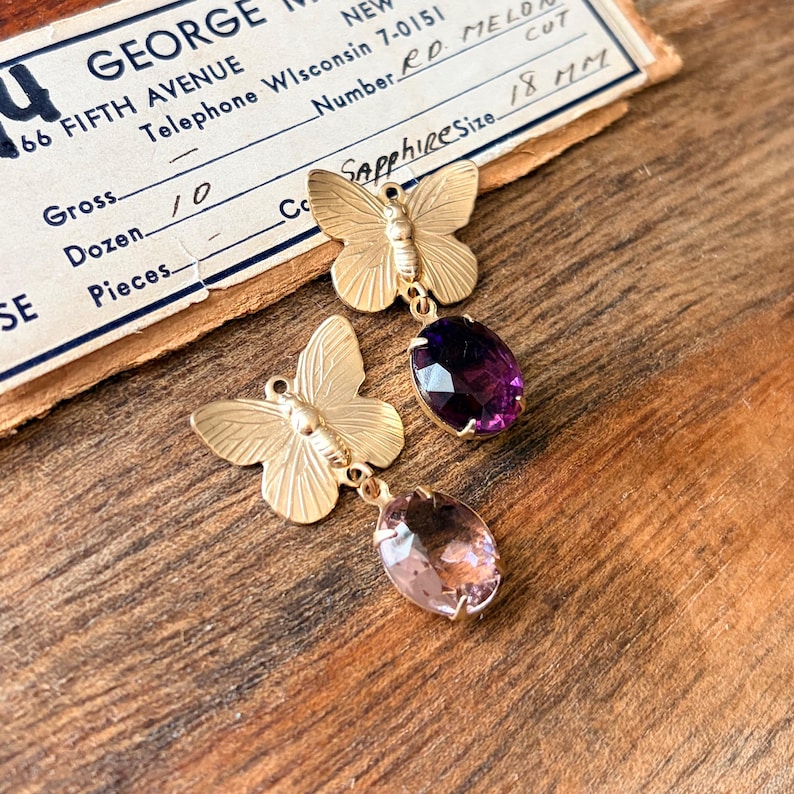 Pu&ograve; includere: Orecchini a farfalla color oro con pietre preziose sfaccettate viola e rosa. Gli orecchini sono esposti su una superficie di legno con un biglietto da visita vintage sullo sfondo. La carta recita "Sapphire Size 18 MM".