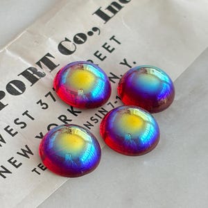 May include: Four round, dome-shaped cabochons featuring an iridescent finish that shifts between vibrant yellow, magenta, and blue hues. The cabochons are arranged on a piece of vintage paper with printed text reading PORT CO., INC. WEST 37TH STREET, NEW YORK, N.Y.