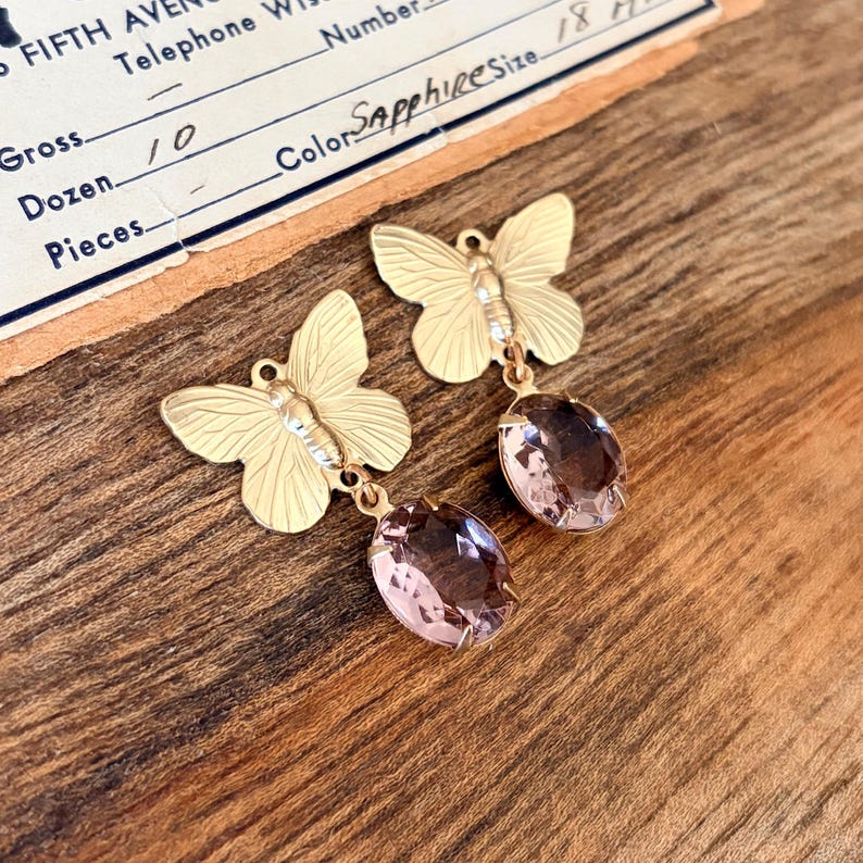 Pu&ograve; includere: Un paio di orecchini a farfalla color oro con gocce di gemme rosa ovali. Gli orecchini sono appoggiati su una superficie di legno, con una fattura vintage visibile sullo sfondo. La fattura recita "Sapphire Size 18".