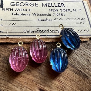 May include: Four vintage gemstone pendants in shades of pink and blue, with gold-colored metal settings. The pendants have an oval, ribbed design. A vintage business card is in the background, with text that reads "GEORGE MELLER, FIFTH AVENUE, NEW YORK, N.Y."
