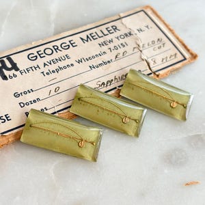 May include: Three rectangular, translucent olive-green buttons featuring a gold-colored fishing rod graphic. The buttons are positioned on a vintage paper label printed with the text GEORGE MELLER, NEW YORK, N.Y., 66 FIFTH AVENUE, and SAPPHIRE.
