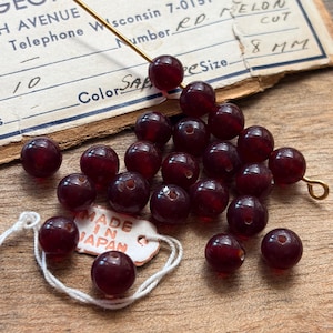 May include: A collection of round, deep red translucent beads with a smooth finish. Some beads are strung on a thin gold wire, while others are loose. A small white tag with the text MADE IN JAPAN is visible, along with a vintage paper label.
