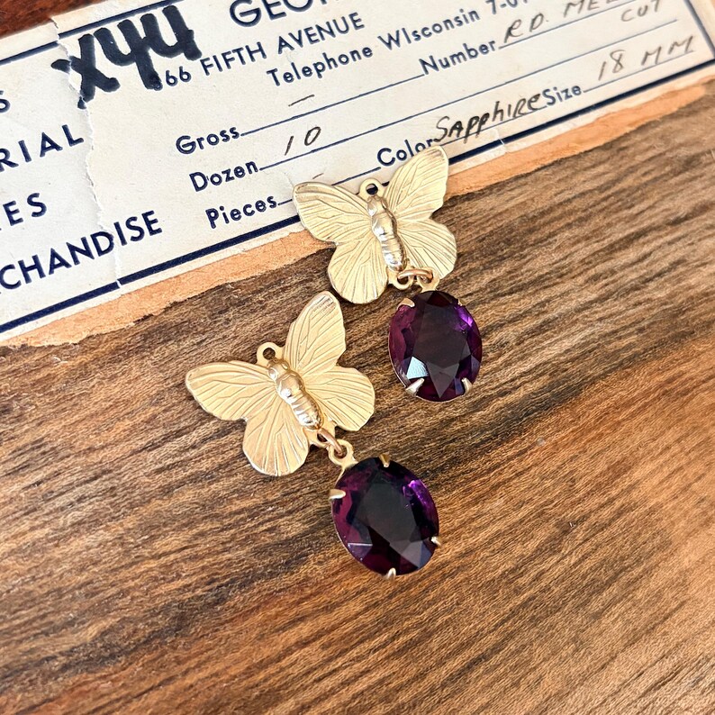 Pu&ograve; includere: Orecchini a farfalla color oro con gemme ovali viola scuro. Gli orecchini sono esposti su una superficie di legno con un documento commerciale vintage sullo sfondo. Il documento include testo come "166 FIFTH AVENUE" e "Sapphire Size 18 mm".