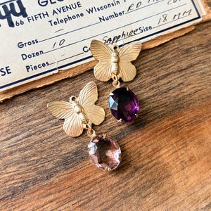 Pu&ograve; includere: Orecchini a farfalla color oro con gemme ovali. Un orecchino presenta una gemma viola scuro, l'altro rosa chiaro. Gli orecchini sono esposti su una superficie di legno con un documento vintage sullo sfondo. Sapphire Size 18 mm.