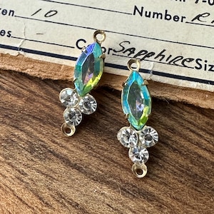 May include: Two vintage gold-toned earring components. Each features an iridescent marquise-shaped crystal above a cluster of clear, round crystals. The components have small loops for attachment. The background includes a piece of aged paper with handwritten text.