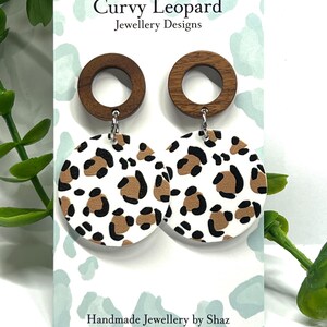 May include: A pair of dangle earrings with a white background and brown and black leopard print. The earrings have a wooden circle at the top and a silver hook.