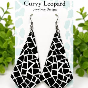 Op de afbeelding: Een paar hangende oorbellen met een geometrisch mozaïekpatroon van zwarte en witte vormen. De oorbellen worden gepresenteerd op een witte kaart met de tekst Curvy Leopard Jewellery Designs en Handmade Jewellery by Saz erop gedrukt.