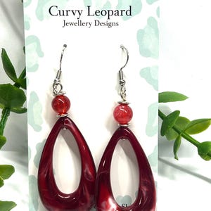 Puede incluir: Pendientes rojos en forma de lágrima con diseño veteado blanco. Cada pendiente tiene una cuenta roja y un gancho plateado. Se exhiben en una tarjeta blanca con el texto "Curvy Leopard Jewellery Designs".