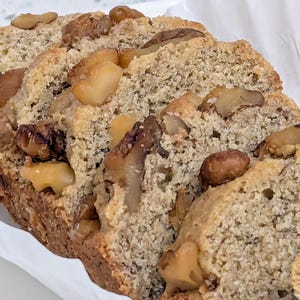May include: Close-up of sliced banana and walnut bread. The bread has a golden-brown crust and a soft, moist interior. Walnuts are scattered on top, adding texture and flavour. The bread is presented on a white surface.
