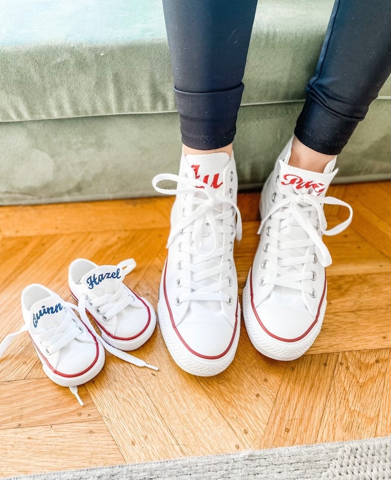 Puede incluir: Un par de zapatillas blancas de ca&ntilde;a alta con texto bordado en rojo, "Lulu" y "Puki", y dos zapatillas blancas m&aacute;s peque&ntilde;as con texto bordado en azul, "Hazel" y "Quinn".