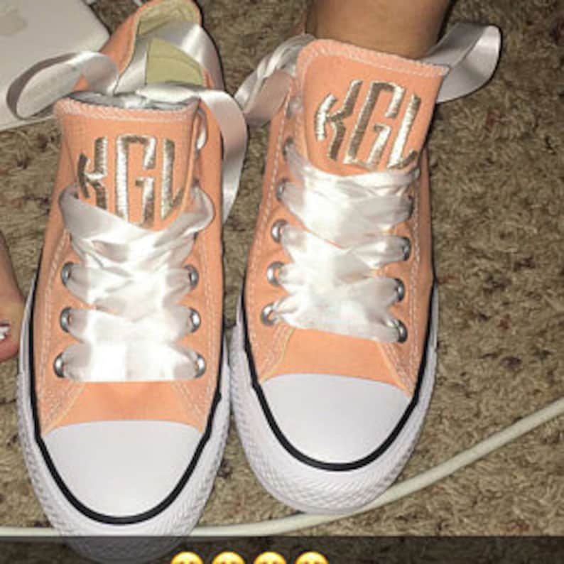 Puede incluir: Un par de zapatillas de lona de color melocot&oacute;n con cordones blancos y suelas blancas. Las zapatillas tienen un monograma dorado "KGL" bordado en el lateral.