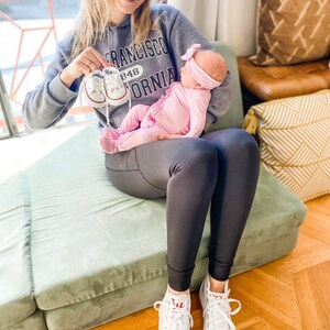 Puede incluir: Una mujer con una sudadera gris con la frase "San Francisco 848 California" impresa en ella, sostiene a un beb&eacute; que lleva un atuendo rosa y una diadema rosa. La mujer sostiene un par de zapatos de beb&eacute; blancos con la palabra "Hard" escrita en ellos. La mujer est&aacute; sentada en un coj&iacute;n verde.