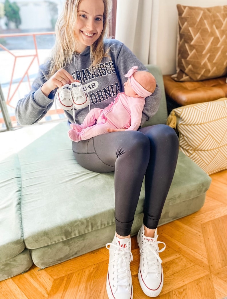 Puede incluir: Una mujer con una sudadera gris con la frase "San Francisco 848 California" impresa en ella, sostiene a un beb&eacute; que lleva un atuendo rosa y una diadema rosa. La mujer sostiene un par de zapatos de beb&eacute; blancos con la palabra "Hard" escrita en ellos. La mujer est&aacute; sentada en un coj&iacute;n verde.