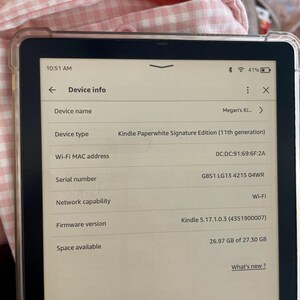 Op de afbeelding: Een Kindle Paperwhite Signature Edition e-reader met een witte voorkant en zwarte randen. Het scherm toont apparaatgegevens, waaronder de apparaatnaam "Megan's Ki...", het apparaattype "Kindle Paperwhite Signature Edition (11th generation)", het Wi-Fi MAC-adres, het serienummer, de netwerkmogelijkheid, de firmwareversie en de beschikbare opslagruimte.