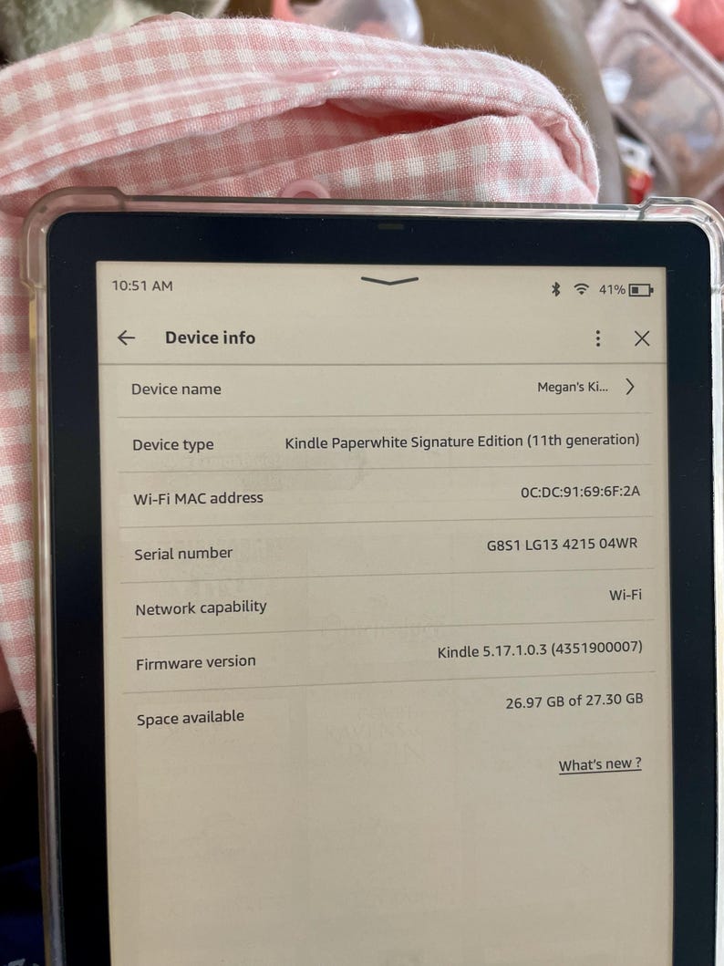 Op de afbeelding: Een Kindle Paperwhite Signature Edition e-reader met een witte voorkant en zwarte randen. Het scherm toont apparaatgegevens, waaronder de apparaatnaam "Megan's Ki...", het apparaattype "Kindle Paperwhite Signature Edition (11th generation)", het Wi-Fi MAC-adres, het serienummer, de netwerkmogelijkheid, de firmwareversie en de beschikbare opslagruimte.