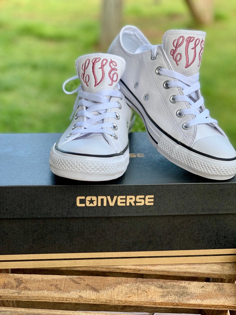 Puede incluir: Un par de zapatillas Converse de cuero blanco con las iniciales bordadas en rosa "LFF" en el lateral. Las zapatillas est&aacute;n en una caja blanca con la palabra "CONVERSE" impresa en la parte superior.
