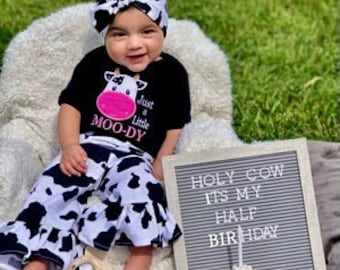 May include: A baby girl wearing a black and white cow print outfit with a pink cow face on the shirt. The shirt says "Just a Little Moo-dy". The baby is sitting on a chair with a sign that says "Holy Cow It's My Half Birthday".