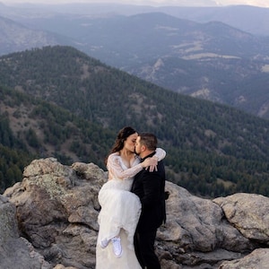 Puede incluir: Una novia y un novio se besan en la cima de una monta&ntilde;a con vistas a colinas onduladas en el fondo. La novia lleva un vestido de novia blanco y el novio lleva un traje negro. La novia est&aacute; en los brazos del novio.