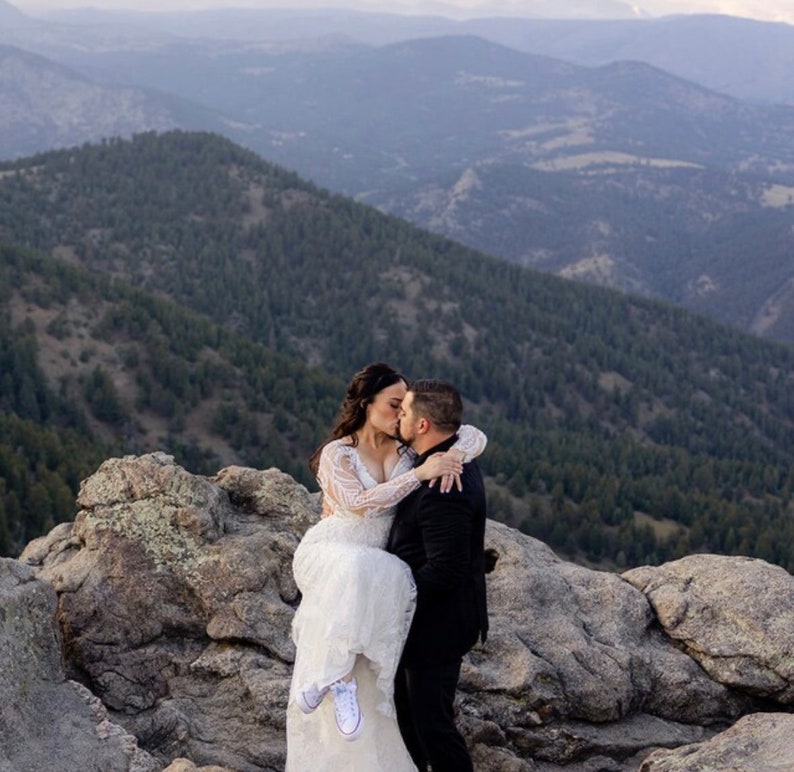 Puede incluir: Una novia y un novio se besan en la cima de una monta&ntilde;a con vistas a colinas onduladas en el fondo. La novia lleva un vestido de novia blanco y el novio lleva un traje negro. La novia est&aacute; en los brazos del novio.