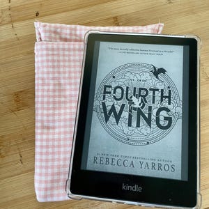 Op de afbeelding: Een Kindle e-reader in een roze en witte geruite hoes. Het scherm toont de cover van het boek "Fourth Wing" van Rebecca Yarros. De boekomslag heeft een zwart-witte illustratie van een draak en de tekst "Fly...or Die".