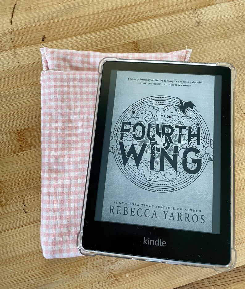 Op de afbeelding: Een Kindle e-reader in een roze en witte geruite hoes. Het scherm toont de cover van het boek "Fourth Wing" van Rebecca Yarros. De boekomslag heeft een zwart-witte illustratie van een draak en de tekst "Fly...or Die".