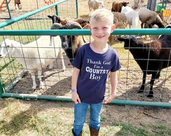 May include: A young boy wearing a navy blue t-shirt with the text "Thank God I'm a Country Boy" in white lettering, stands in front of a fence with sheep and goats. He is wearing blue jeans and brown cowboy boots.