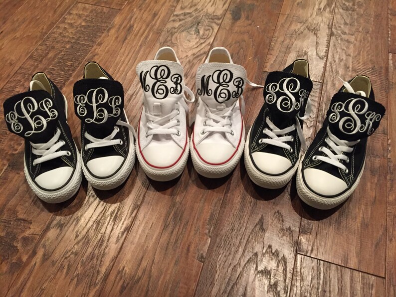 Puede incluir: Seis pares de zapatos de lona negros y blancos con solapas de tela negra que tienen iniciales bordadas en blanco. Las iniciales son "EB", "LL", "EB", "MB", "SS" y "SS".