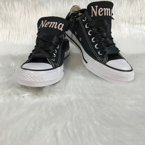 Puede incluir: Un par de zapatillas de lona negras con suelas blancas y cordones negros. Las zapatillas tienen una cinta rosa atada alrededor y el nombre "Nema" est&aacute; bordado en la cinta con hilo rosa.