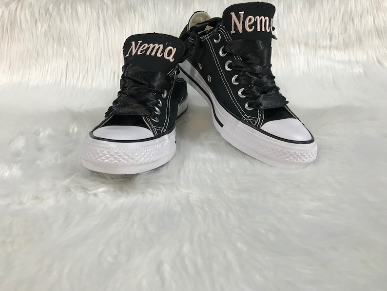 Puede incluir: Un par de zapatillas de lona negras con suelas blancas y cordones negros. Las zapatillas tienen una cinta rosa atada alrededor y el nombre "Nema" est&aacute; bordado en la cinta con hilo rosa.