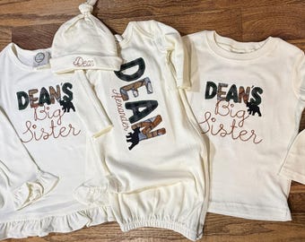 Może przedstawiać: Dwie koszulki z długim rękawem w kolorze złamanej bieli z falbankami u dołu, każda z napisem "DEAN'S Big Sister" wyhaftowanym na czarno i brązowo z sylwetką niedźwiedzia. Pasująca koszula nocna dla niemowląt i czapeczka z węzłem z haftem "Dean".
