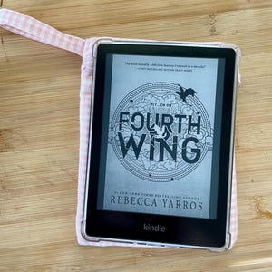 Op de afbeelding: Een zwart-witte Kindle e-reader met een roze en witte gingham hoes. Het scherm toont de cover van het boek "Fourth Wing" van Rebecca Yarros. De boekomslag heeft een zwart-witte illustratie van een draak en de tekst "Fly...or Die."