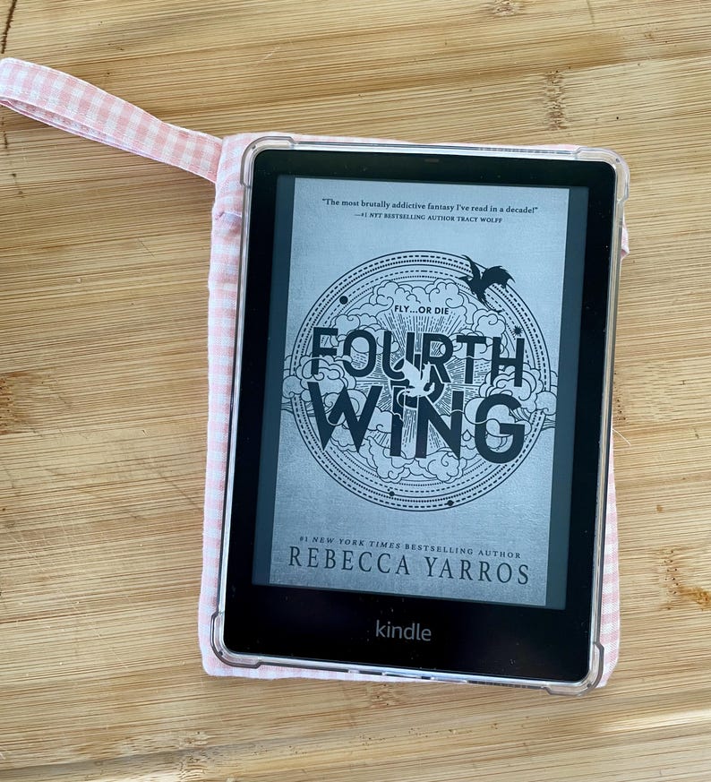 Op de afbeelding: Een zwart-witte Kindle e-reader met een roze en witte gingham hoes. Het scherm toont de cover van het boek "Fourth Wing" van Rebecca Yarros. De boekomslag heeft een zwart-witte illustratie van een draak en de tekst "Fly...or Die."