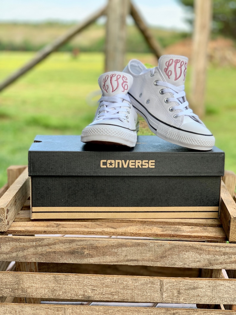 Puede incluir: Un par de zapatillas blancas de lona con iniciales bordadas en rosa en el lateral. Las zapatillas est&aacute;n encima de una caja de zapatos Converse negra.