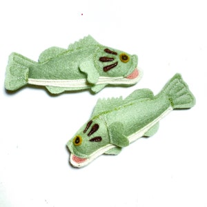 May include: Two handmade felt fish in a light green color. The fish have brown embroidered details for the eyes and gills, with a white belly and pink mouth. The fish are designed with a whimsical, cartoonish style.