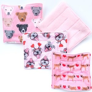 May include: Four pink and white fabric pot holders with a dog theme. One pot holder has a repeating pattern of dogs with hearts and stripes. Another pot holder has a repeating pattern of dogs with hearts. The other two pot holders are solid pink.