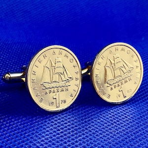 Può includere: Gemelli dorati con un design a moneta raffigurante una nave a vela. Le monete presentano testo greco e gli anni 1978 e 1982. I gemelli sono su sfondo blu.