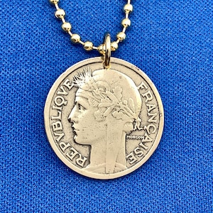 以下が含まれることがあります： 金のチェーンネックレスに、金の硬貨のペンダントがぶら下がっています。ペンダントには、月桂樹の冠をかぶった女性の横顔が描かれています。硬貨の縁には「REPUBLIQUE FRANCAISE」という文字が刻まれています。