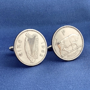 May include: Silver cufflinks featuring Irish coins. One cufflink displays a harp and the year 1962, while the other shows a bull and the inscription "1s scilling". The cufflinks are set against a blue background.