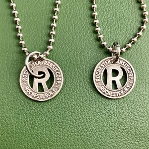 Könnte beinhalten: Zwei silberfarbene Kugelkettchen aus Metall, jeweils mit einem runden Anhänger, der in der Mitte ein ausgeschnittenes R aufweist. Der Text Rochester Transit Corp N.Y. ist am Rand jedes runden Anhängers eingeprägt.