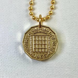 May include: A gold-colored necklace featuring a pendant with a vintage British three pence coin from 1966. The coin pendant has a crown and a grid design. The necklace chain is made of small, round beads.