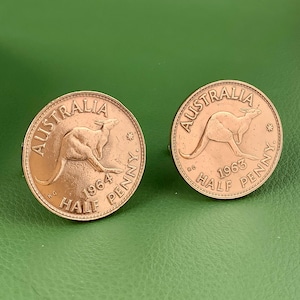 May include: Two copper-colored cufflinks featuring Australian half-penny coins. Each coin displays a kangaroo and the words "Australia" and "Half Penny", with the years 1963 and 1964. The cufflinks are set against a green background.