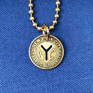 May include: A gold-coloured pendant necklace. The circular charm is inscribed with "NEW YORK CITY TRANSIT AUTHORITY" around the edge. The centre of the charm displays the letters "NYC". The necklace has a ball chain.