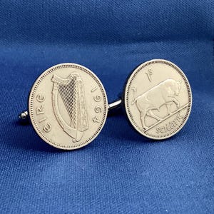May include: Silver cufflinks featuring Irish coins. One cufflink displays a harp and the word "Eire" with the year 1964. The other cufflink shows a bull and the word "scilling". The cufflinks are set against a blue background.