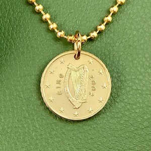 May include: A gold coin pendant necklace. The pendant displays a harp design, the word "eire", and the year "2002". The necklace has a gold ball chain. The background is green.