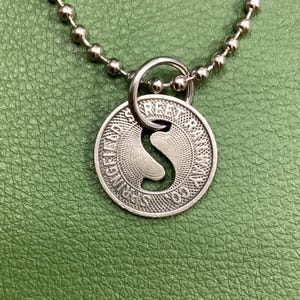 Könnte beinhalten: Silberfarbene Anhängerkette mit einem kreisförmigen Anhänger mit geprägtem Design und dem Text "Springs Free". Der Anhänger hat eine Aussparung. Die Halskette hat eine Kugelkette und einen Ringverschluss.