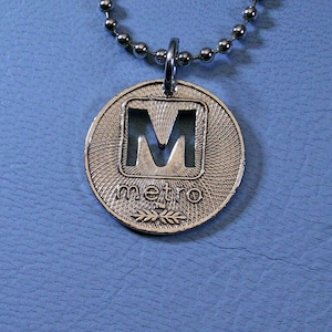 May include: Silver necklace with a round pendant featuring a cutout letter "M" inside a square and the word "metro" below.