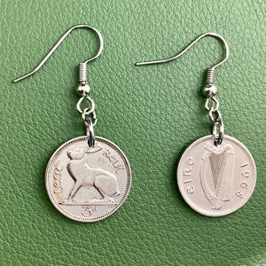 May include: Silver-toned earrings featuring coin pendants. One earring displays a hare, the other a harp, both with text. The earrings hang from silver-colored fish hook ear wires. The background is a solid green.